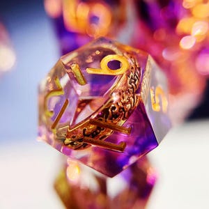 May include: Close-up of a polyhedral die with a purple and gold color scheme. The die has gold numerals and intricate gold detailing inside the clear resin. The background is blurred with hints of purple and orange.