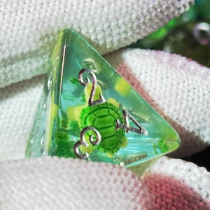 May include: Close-up of a set of polyhedral dice. The focus is on a green and yellow D4 die with a turtle design. The die is held in a white glove. Other dice are visible in the background, with a mix of green, blue, and purple hues.