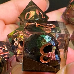 May include: A set of multi-sided dice with a dark, shimmering appearance. The dice have copper-coloured numerals and some feature a skull design. Ideal for tabletop gaming.