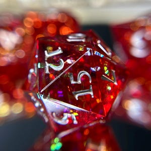 May include: A close-up of a red, multi-sided die with silver numbering. The die is transparent, with iridescent flecks visible inside. Other dice are blurred in the background, creating a bokeh effect. This is a gaming accessory.
