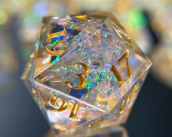 Peut inclure: Un dé D20 clair et à multiples facettes avec des chiffres dorés. Le dé est rempli de paillettes irisées, créant un effet chatoyant. Les chiffres sont clairement visibles, et le dé est posé sur un fond flou.