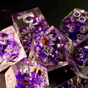 Könnte beinhalten: Ein Set aus sieben polyedrischen Würfeln. Jeder Würfel besteht aus klarem Harz, gefüllt mit lila Blumen und silbernen Zahlen. Die Würfel haben verschiedene Formen, darunter ein d4, d6, d8, d10, d12, d20 und ein Prozentwürfel.
