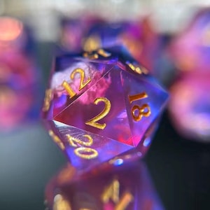 Puede incluir: Primer plano de un dado de veinte caras con un diseño veteado púrpura y rosa. Los números son dorados. El dado está enfocado, con otros dados desenfocados en el fondo. El dado se refleja en una superficie oscura.