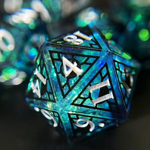 Puede incluir: Primer plano de un conjunto de dados poliédricos, con un enfoque en un d20. Los dados son de color verde azulado y azul con números plateados. El d20 tiene un diseño de vidriera con líneas negras. Otros dados están borrosos en el fondo.