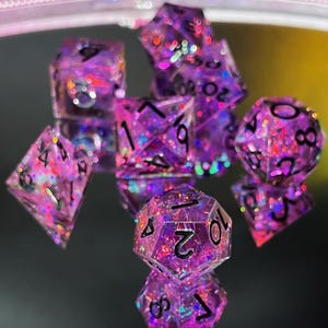 Puede incluir: Un juego de siete dados de purpurina morados y rosas con números negros. Los dados tienen varias formas, incluyendo un d4, d6, d8, d10, d12, d20 y un dado de porcentaje. Los dados son reflectantes y parecen estar hechos de resina.