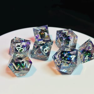 Op de afbeelding: Een set van zeven iriserende dobbelstenen met witte nummers. De dobbelstenen zijn gemaakt van helder hars met ingebedde glitter en kleurwervelingen. De dobbelstenen zijn in verschillende vormen, waaronder een d4, d6, d8, d10, d12 en d20.