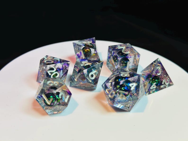 Op de afbeelding: Een set van zeven iriserende dobbelstenen met witte nummers. De dobbelstenen zijn gemaakt van helder hars met ingebedde glitter en kleurwervelingen. De dobbelstenen zijn in verschillende vormen, waaronder een d4, d6, d8, d10, d12 en d20.