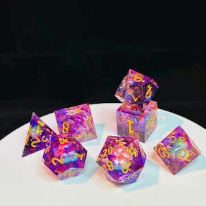 Op de afbeelding: Een set van zeven regenboogkleurige paarse en roze dobbelstenen met gouden cijfers. De dobbelstenen liggen op een wit oppervlak. De dobbelstenen zijn in verschillende vormen, waaronder een d4, d6, d8, d10, d12 en d20.