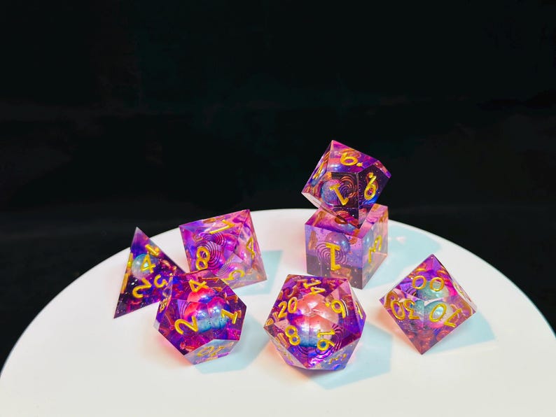 Op de afbeelding: Een set van zeven regenboogkleurige paarse en roze dobbelstenen met gouden cijfers. De dobbelstenen liggen op een wit oppervlak. De dobbelstenen zijn in verschillende vormen, waaronder een d4, d6, d8, d10, d12 en d20.