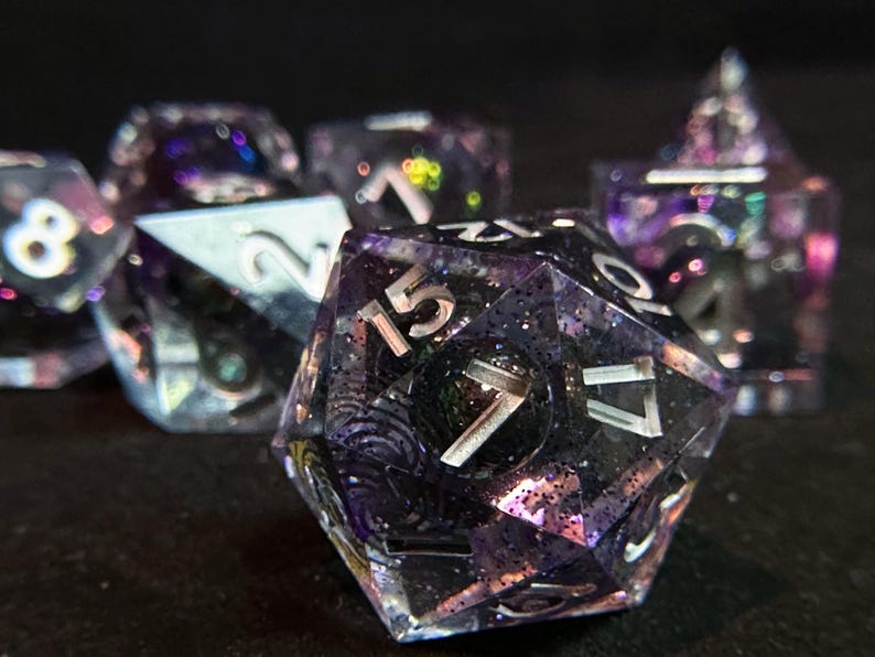 Op de afbeelding: Een set iriserende dobbelstenen met zilveren nummers. De dobbelstenen zijn gemaakt van helder hars met een paars en zwart swirl-patroon. De dobbelstenen zijn perfect voor tabletop gaming.