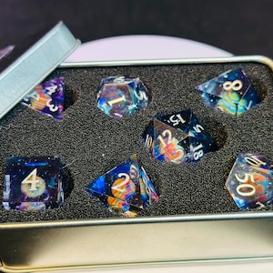 Op de afbeelding: Een set van zeven iriserende dobbelstenen met een galaxie thema. De dobbelstenen zitten in een zilveren blik met zwarte schuimvulling. De dobbelstenen zijn genummerd van 1 tot 6 en 10, 12 en 18.