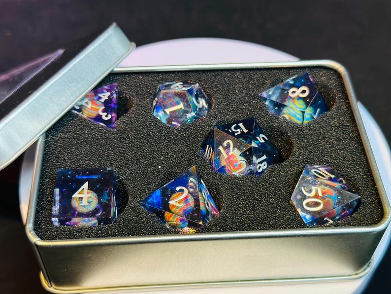 Op de afbeelding: Een set van zeven iriserende dobbelstenen met een galaxie thema. De dobbelstenen zitten in een zilveren blik met zwarte schuimvulling. De dobbelstenen zijn genummerd van 1 tot 6 en 10, 12 en 18.