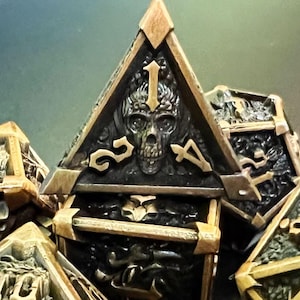 May include: Close-up of a set of polyhedral dice with a gothic theme. The dice are black with gold-colored trim and feature skull and number designs. The dice are arranged in a pile, with the skull-faced die at the top.