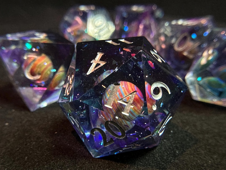 Op de afbeelding: Een set van zes iriserende dobbelstenen met een galactisch patroon. De dobbelstenen zijn gemaakt van helder hars met ingebedde glitter en holografische vlokken. De nummers op de dobbelstenen zijn zilverkleurig en zichtbaar door het hars. De dobbelstenen zijn scherp in beeld, de achtergrond is wazig.
