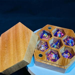 Op de afbeelding: Een zeshoekige houten doos met een deksel, met daarin zeven iriserende dobbelstenen met gouden cijfers. De dobbelstenen zijn paars en roze met gouden spikkels.