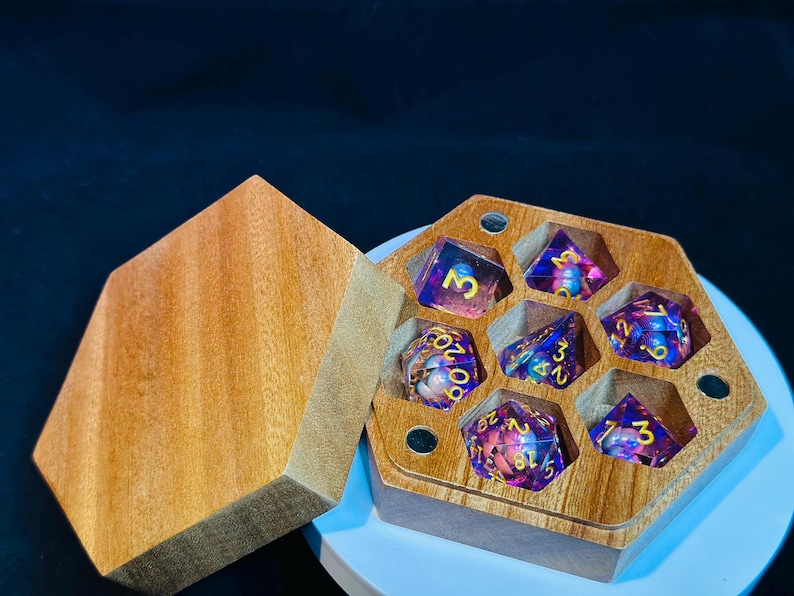 Op de afbeelding: Een zeshoekige houten doos met een deksel, met daarin zeven iriserende dobbelstenen met gouden cijfers. De dobbelstenen zijn paars en roze met gouden spikkels.