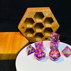 Op de afbeelding: Een set van iriserende paarse en gouden dobbelstenen met gouden nummers, in een houten zeshoekige doos met een deksel. De dobbelstenen liggen op een wit oppervlak.