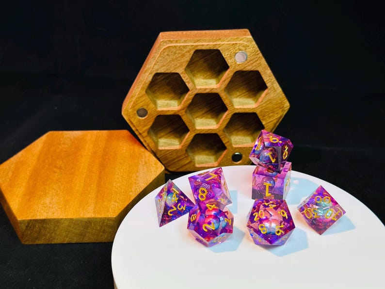 Op de afbeelding: Een set van iriserende paarse en gouden dobbelstenen met gouden nummers, in een houten zeshoekige doos met een deksel. De dobbelstenen liggen op een wit oppervlak.