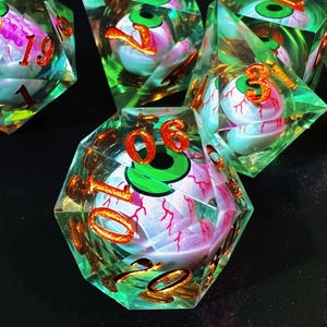 May include: A set of polyhedral dice with a spooky theme. Each die is clear with a green and pink eyeball design inside. The numbers are in a metallic orange color, and the dice are set against a dark background.