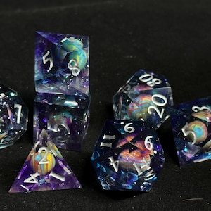 Op de afbeelding: Een set van zeven polyhedrische dobbelstenen met een galaxie thema. De dobbelstenen zijn helder met een draaiende blauwe en paarse achtergrond en zilveren glitter. Elke dobbelsteen heeft een planeet van een andere kleur ingebed in de hars. De nummers op de dobbelstenen zijn wit.