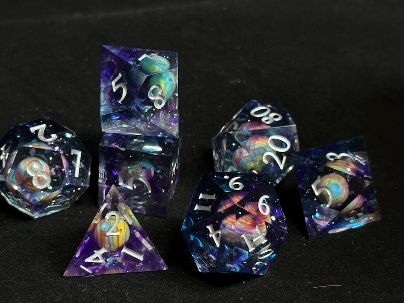Op de afbeelding: Een set van zeven polyhedrische dobbelstenen met een galaxie thema. De dobbelstenen zijn helder met een draaiende blauwe en paarse achtergrond en zilveren glitter. Elke dobbelsteen heeft een planeet van een andere kleur ingebed in de hars. De nummers op de dobbelstenen zijn wit.