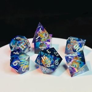 Op de afbeelding: Set van zeven polyhedrische dobbelstenen met een galaxie thema. De dobbelstenen zijn helder met blauwe, paarse en roze swirls en glitter. De nummers zijn wit en zwart omrand. De dobbelstenen liggen op een wit oppervlak.