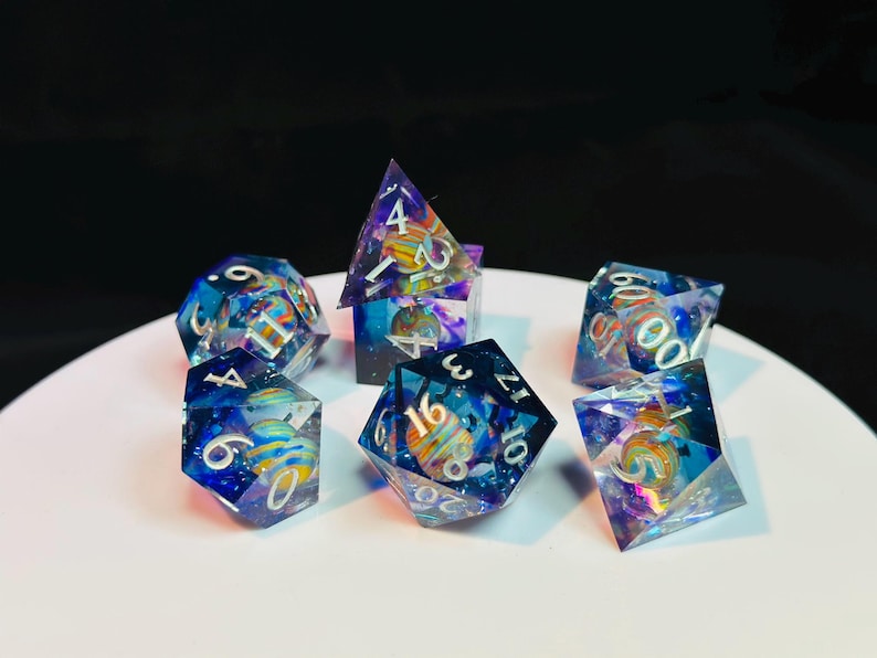 Op de afbeelding: Set van zeven polyhedrische dobbelstenen met een galaxie thema. De dobbelstenen zijn helder met blauwe, paarse en roze swirls en glitter. De nummers zijn wit en zwart omrand. De dobbelstenen liggen op een wit oppervlak.
