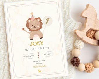 May include: A birthday invitation with a lion illustration, the text "JOEY IS TURNING ONE", and event details. The invitation is cream-colored with a gold border. A wooden toy and a beaded teether are next to the invitation.