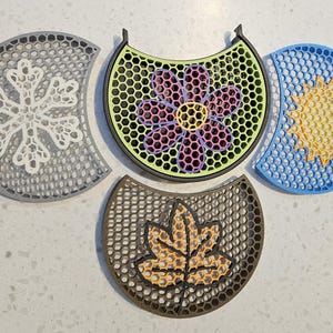 May include: Four decorative coasters with seasonal designs. A gray snowflake, a flower with pink and purple petals, a yellow sun, and a brown leaf. Each coaster has a honeycomb pattern.