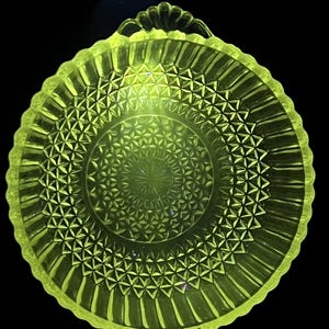 Puede incluir: Un plato redondo de vidrio amarillo fluorescente con un patrón geométrico prensado detallado. El centro muestra un diseño de estrella rodeado por anillos concéntricos de pequeños diamantes, mientras que el borde exterior tiene estrías verticales.