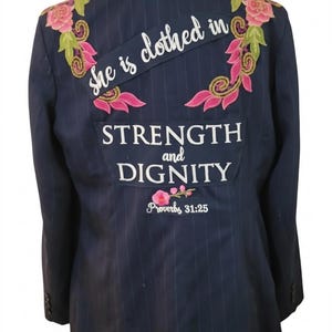 May include: Navy blue pinstripe blazer with a denim collar. The back features embroidered pink and green floral accents and the text "She is clothed in STRENGTH and DIGNITY Proverbs 31:25."