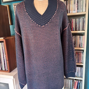 May include: A navy blue sweater with a V-neck collar and long sleeves. The sweater has a textured pattern in navy and peach. The collar and seams are accented with peach stitching. The sweater is displayed on a mannequin.