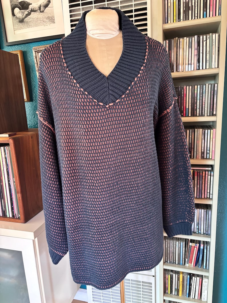 May include: A navy blue sweater with a V-neck collar and long sleeves. The sweater has a textured pattern in navy and peach. The collar and seams are accented with peach stitching. The sweater is displayed on a mannequin.