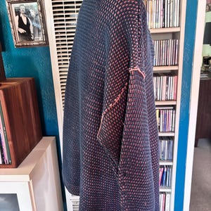 May include: A navy blue and coral-colored knit sweater with a high collar. The sweater has a textured pattern and coral-colored stitching along the seams. The sweater is displayed on a wooden stand.