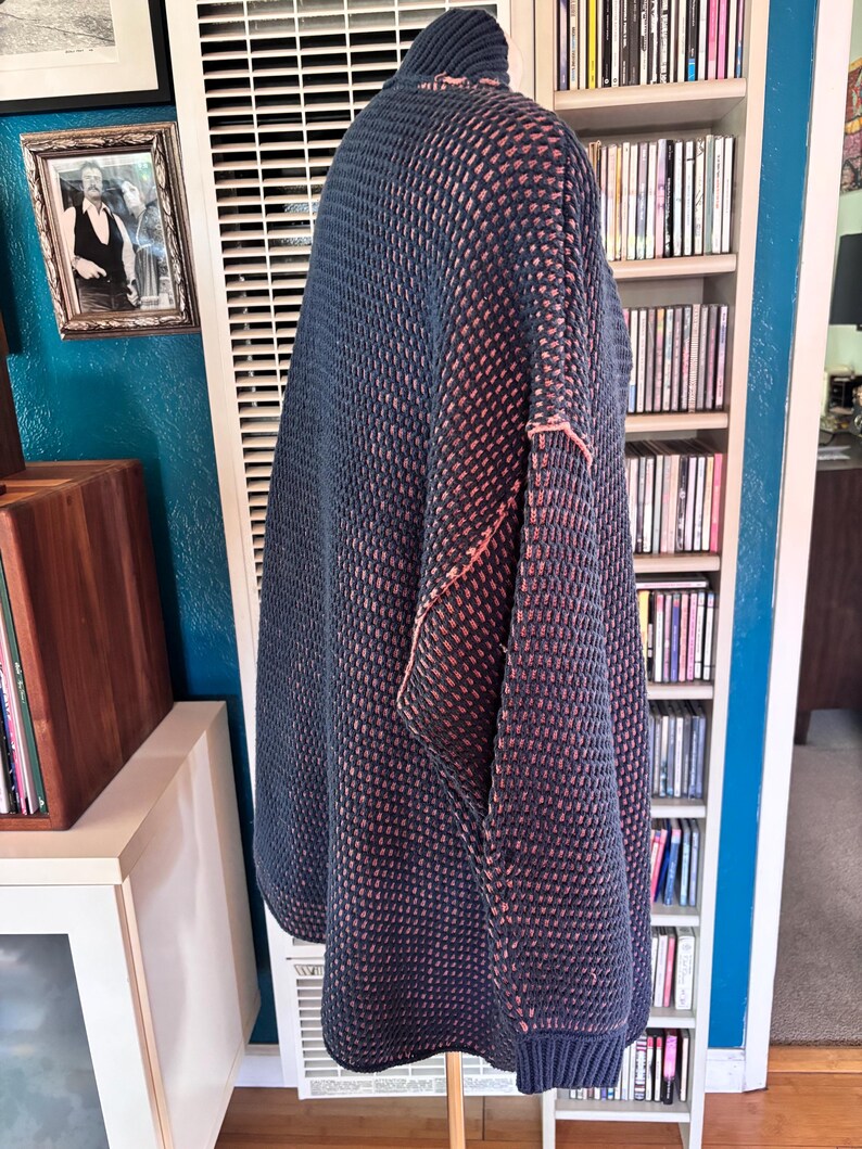 May include: A navy blue and coral-colored knit sweater with a high collar. The sweater has a textured pattern and coral-colored stitching along the seams. The sweater is displayed on a wooden stand.
