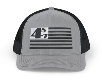Pode incluir: Um chapéu de caminhoneiro cinza e preto com um design de bandeira americana em preto e branco e o texto "45-7" na frente.