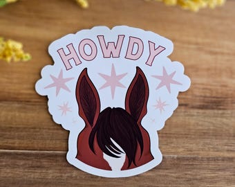 以下が含まれることがあります： ピンクと白の馬の頭のイラストとピンクの文字で「HOWDY」と書かれた白いステッカー。馬の頭は茶色の耳と黒いたてがみをしている。