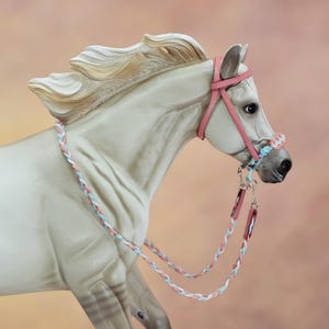 May include: A model horse figurine in a light cream colour with a flowing mane, wearing a pink leather bridle and a braided rein set featuring interwoven light blue, white, and pink cord. The bridle includes a noseband with matching braided accents.