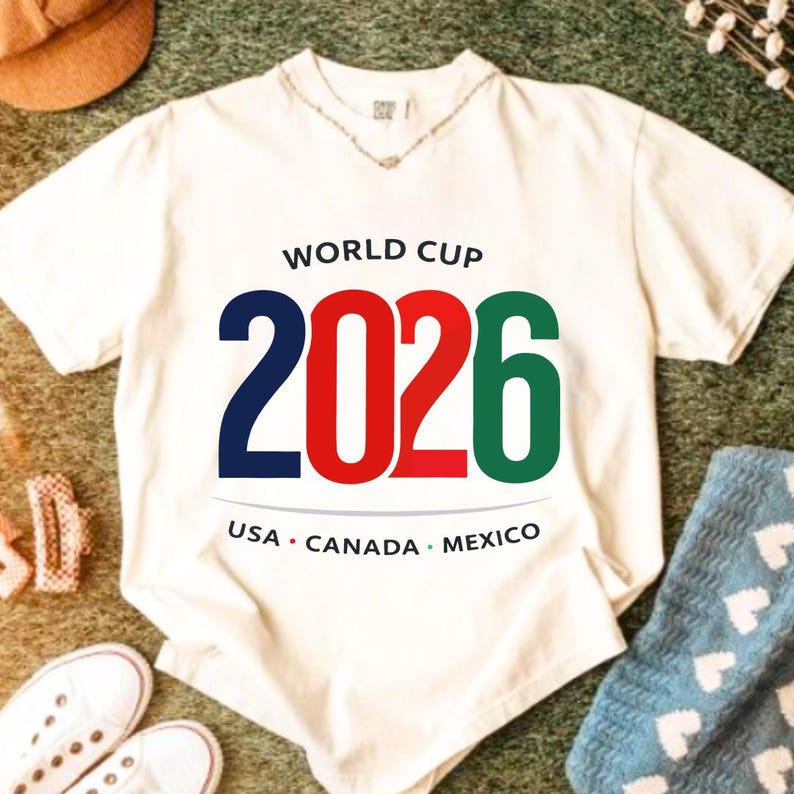 May include: A white t-shirt with the text "WORLD CUP 2026" in navy blue, red, and green. Below the text are the words "USA CANADA MEXICO" in a semi-circle. The shirt is on a green surface with other items in the background.