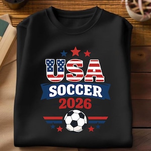 May include: Black crew neck sweatshirt with a graphic design. The design features the text "USA SOCCER 2026" in red, white, and blue, along with a soccer ball and stars. The letters "USA" are designed with the American flag.