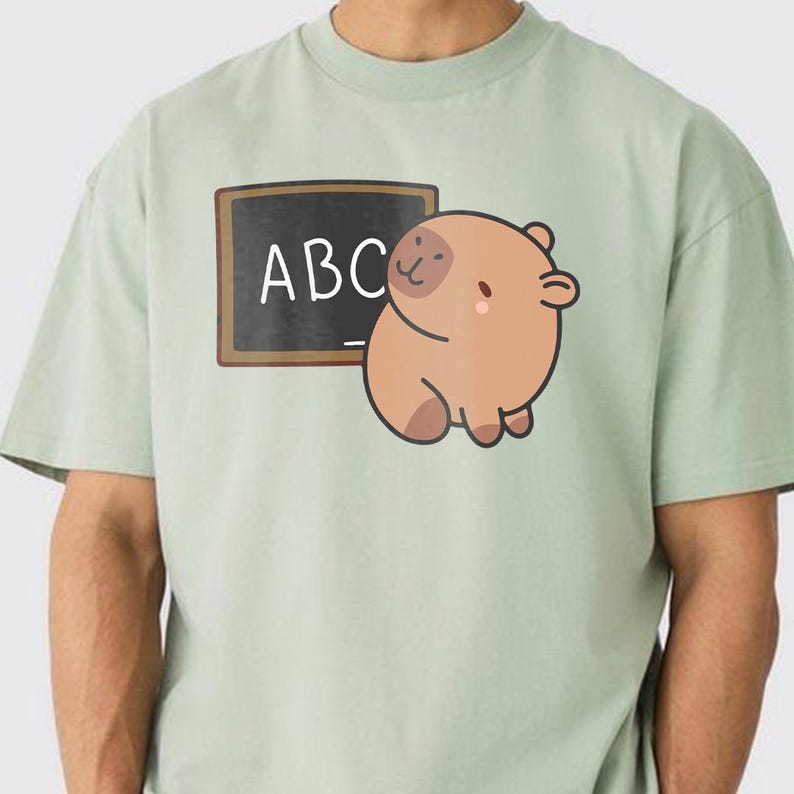 May include: A pale green t-shirt with a cartoon illustration of a brown animal and a chalkboard with the letters "ABC". The t-shirt is made of a soft material. The animal has a pink blush.
