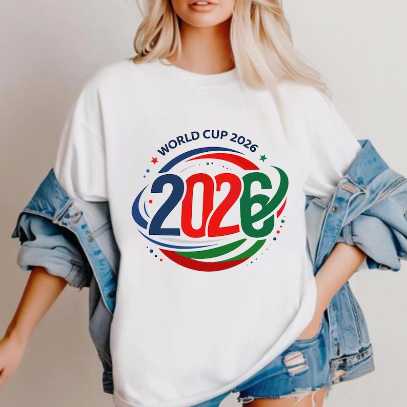 May include: White t-shirt featuring a colorful graphic design for the 2026 World Cup. The design includes the text "WORLD CUP 2026" in a circular arrangement above the number "2026" in blue, red, and green.