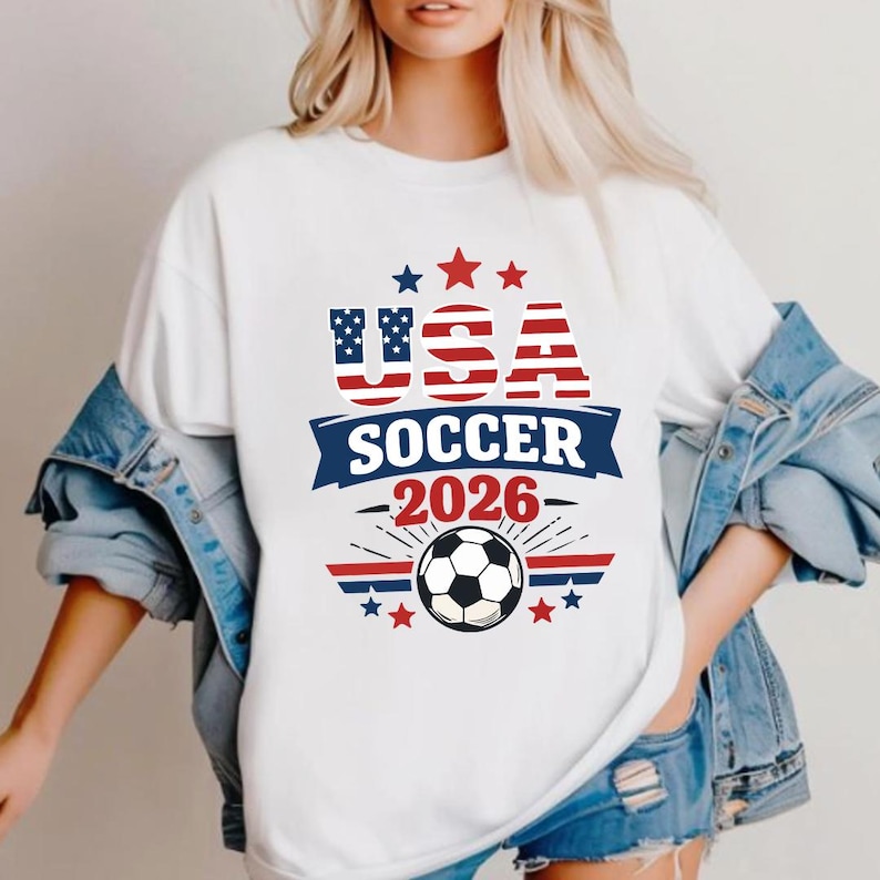 May include: White t-shirt with a graphic design featuring the text "USA SOCCER 2026" in red, white, and blue. The design includes stars, stripes, and a soccer ball. The shirt is a casual wear item.