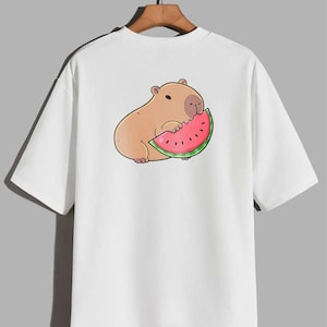 May include: White t-shirt with a cartoon capybara enjoying watermelon. The capybara is light brown with a dark outline, and the watermelon slice is red with black seeds and a green rind. The shirt has short sleeves and a relaxed fit.