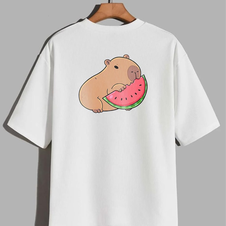 May include: White t-shirt with a cartoon capybara enjoying watermelon. The capybara is light brown with a dark outline, and the watermelon slice is red with black seeds and a green rind. The shirt has short sleeves and a relaxed fit.