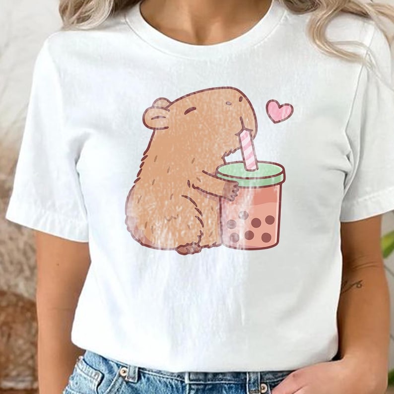 May include: White t-shirt with a cartoon capybara enjoying boba tea. The capybara is brown and holding a pink and green boba tea cup. A pink heart floats above. This casual t-shirt is suitable for everyday wear.