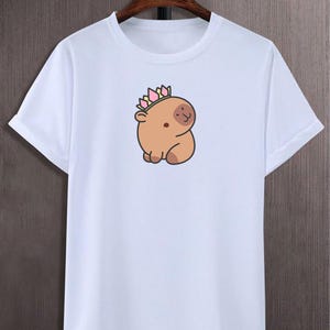 May include: White t-shirt with a cartoon capybara wearing a crown. The capybara is light brown with a small, smiling face and a crown with pink, yellow, and green accents. The shirt has short sleeves and a crew neck.
