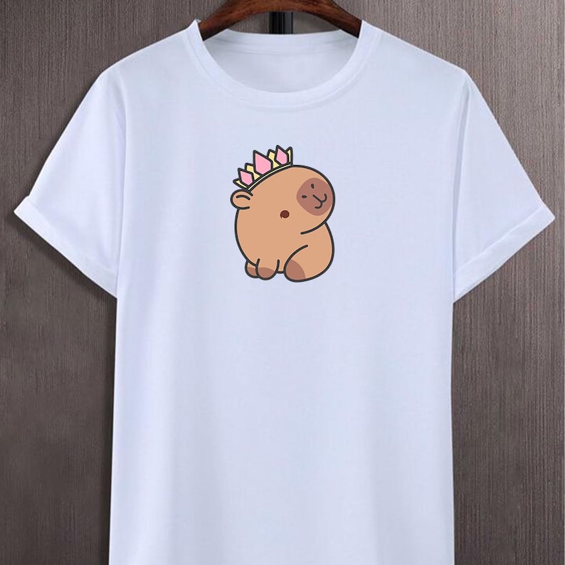 May include: White t-shirt with a cartoon capybara wearing a crown. The capybara is light brown with a small, smiling face and a crown with pink, yellow, and green accents. The shirt has short sleeves and a crew neck.