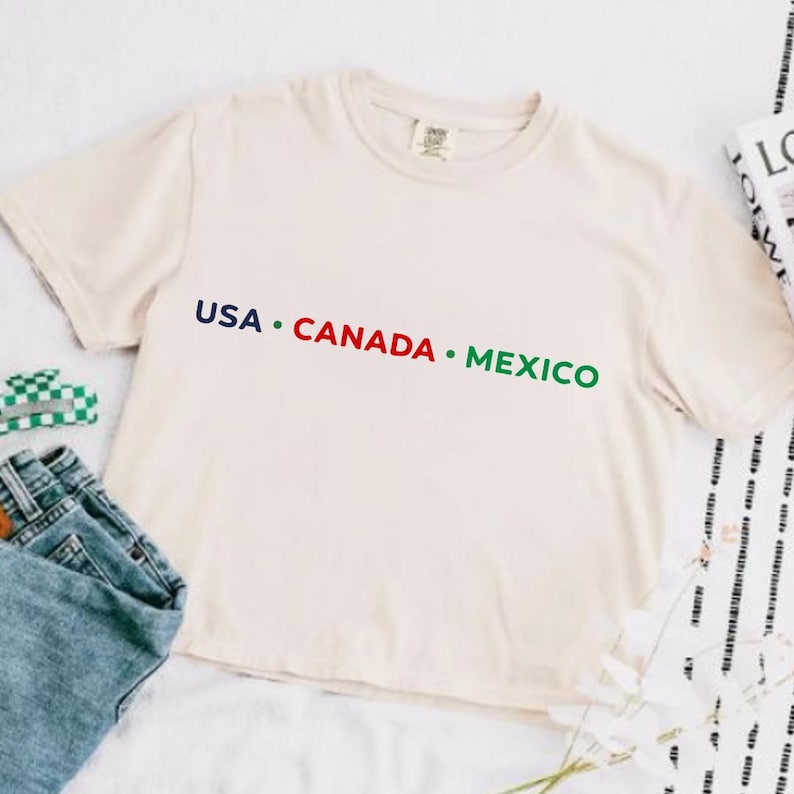 May include: Cream-colored t-shirt with the text "USA &bull; CANADA &bull; MEXICO" in blue, red, and green. The shirt is made of a soft material and has a classic crew neck design. A pair of blue jeans and a small green and white checkered item are visible.