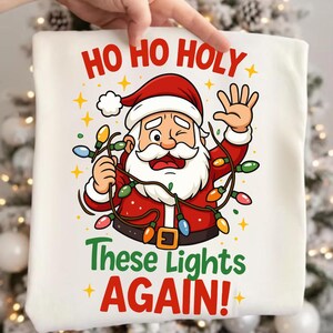 May include: White Christmas-themed sweatshirt featuring a cartoon Santa Claus winking and holding a string of colorful lights. The text "HO HO HOLY" is at the top, with "These Lights AGAIN!" below, all in festive colors.